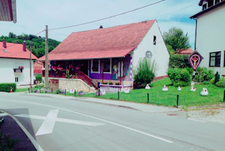 A quaint house with a red-tiled roof and white walls, featuring purple accents and a painted mural on the exterior. The house is adorned with hanging flower pots and surrounded by lush greenery and manicured lawns. The front yard has several decorative items resembling chickens and a small shrine or decorative structure on the side. The road in front of the house shows a large white arrow, and other houses are visible in the background, indicating a residential area.