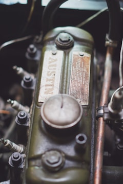 A close-up view of a vintage engine component shows a metal part with a greenish, weathered finish. The surface includes a detailed advisory plate with a prominent bolt atop. Several spark plugs and other mechanical parts surround the main piece, indicating a car engine setup.