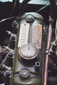 A close-up view of a vintage engine component shows a metal part with a greenish, weathered finish. The surface includes a detailed advisory plate with a prominent bolt atop. Several spark plugs and other mechanical parts surround the main piece, indicating a car engine setup.