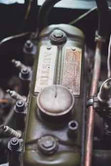 A close-up view of a vintage engine component shows a metal part with a greenish, weathered finish. The surface includes a detailed advisory plate with a prominent bolt atop. Several spark plugs and other mechanical parts surround the main piece, indicating a car engine setup.