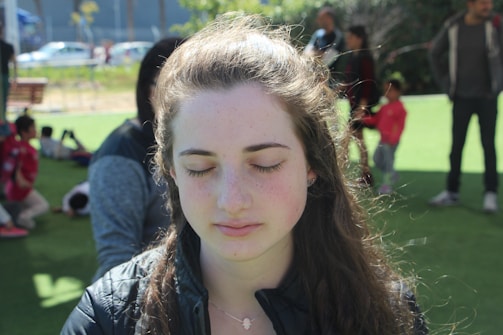 A young woman with long hair is standing outdoors with her eyes closed, appearing calm and relaxed. In the background, people are scattered across a grassy area, with some sitting and others standing. The setting suggests a park-like environment with sunlight filtering through.