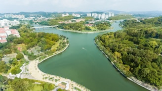 An aerial view of a scenic landscape featuring a winding river surrounded by lush greenery and trees. Residential buildings and structures with red-tiled roofs are visible to the left, while modern high-rise apartments are in the distance. There is a bridge crossing the river in the background, and the area looks like a blend of urban and natural environments.