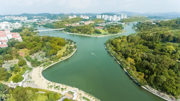 An aerial view of a scenic landscape featuring a winding river surrounded by lush greenery and trees. Residential buildings and structures with red-tiled roofs are visible to the left, while modern high-rise apartments are in the distance. There is a bridge crossing the river in the background, and the area looks like a blend of urban and natural environments.