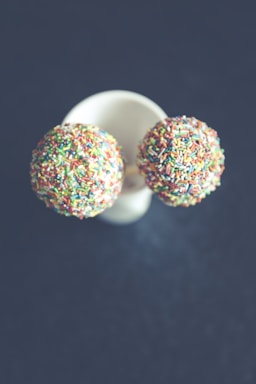Two cake pops covered in colorful sprinkles are placed on a white cup against a dark background. The image is taken from above, emphasizing the contrast between the bright sprinkles and the muted background.
