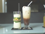 Two tall glasses are placed on a reflective table. The glass on the left contains a creamy drink decorated with a green leaf and a twine bow with small yellow flowers. The glass on the right holds a layered iced coffee drink, topped with frothy cream and a straw. Both are placed on individual black coasters.
