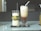 Two tall glasses are placed on a reflective table. The glass on the left contains a creamy drink decorated with a green leaf and a twine bow with small yellow flowers. The glass on the right holds a layered iced coffee drink, topped with frothy cream and a straw. Both are placed on individual black coasters.