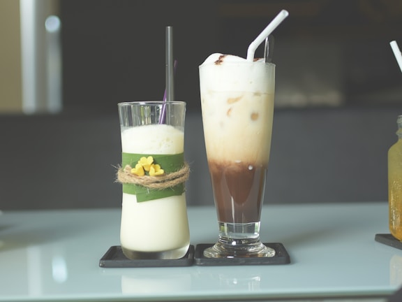 Two tall glasses are placed on a reflective table. The glass on the left contains a creamy drink decorated with a green leaf and a twine bow with small yellow flowers. The glass on the right holds a layered iced coffee drink, topped with frothy cream and a straw. Both are placed on individual black coasters.