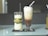 Two tall glasses are placed on a reflective table. The glass on the left contains a creamy drink decorated with a green leaf and a twine bow with small yellow flowers. The glass on the right holds a layered iced coffee drink, topped with frothy cream and a straw. Both are placed on individual black coasters.