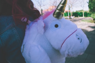 A plush unicorn backpack is being carried on someone's back. The unicorn is white with a golden horn and pink mane.