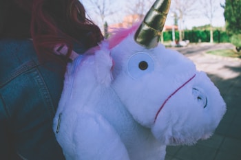 A plush unicorn backpack is being carried on someone's back. The unicorn is white with a golden horn and pink mane.