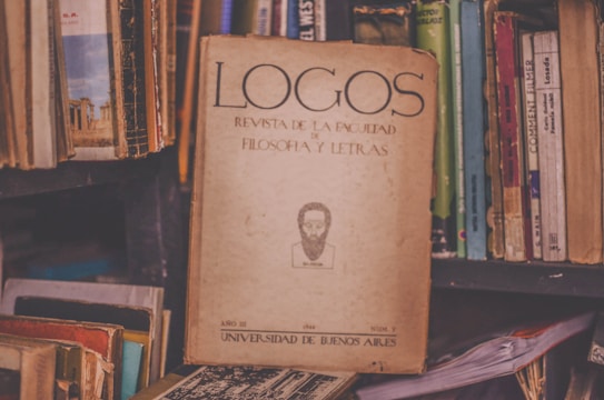 A vintage journal titled 'Logos Revista de la Facultad de Filosof&iacute;a y Letras' is prominently placed among a collection of various books on a cluttered shelf. The worn, yellowed cover contains textual elements and an illustration of a bearded figure. The surrounding books have colorful but faded covers, contributing to a nostalgic and scholarly atmosphere.