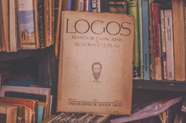A vintage journal titled 'Logos Revista de la Facultad de Filosof&iacute;a y Letras' is prominently placed among a collection of various books on a cluttered shelf. The worn, yellowed cover contains textual elements and an illustration of a bearded figure. The surrounding books have colorful but faded covers, contributing to a nostalgic and scholarly atmosphere.