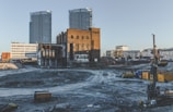 An urban construction site with a mix of old industrial brick buildings and modern skyscrapers in the background. The ground appears excavated, with visible construction machinery and debris. The scene conveys a transitional phase between the old and new architecture.