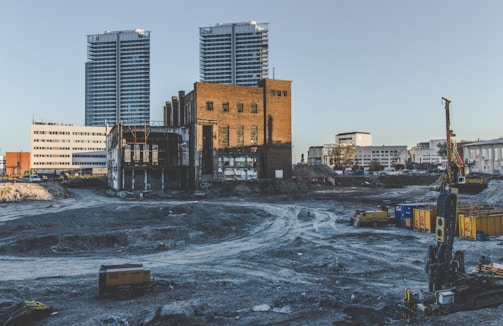 An urban construction site with a mix of old industrial brick buildings and modern skyscrapers in the background. The ground appears excavated, with visible construction machinery and debris. The scene conveys a transitional phase between the old and new architecture.
