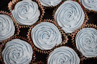 Several cupcakes topped with light blue frosting arranged in a close-up. The frosting is artistically swirled to resemble roses, set in colorful paper liners.