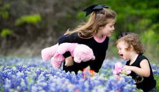 Two young children are playing in a field of blue and white flowers. One child is holding a pink stuffed animal, while the other has a pink object in hand. Both children are dressed in black outfits with large black bows in their hair. They appear to be enjoying their time together, with big smiles and engaging interaction.