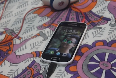 A Samsung smartphone displaying a lock screen with a person's face wearing reflective sunglasses. The phone is placed on a colorful patterned fabric with abstract designs in purple, orange, and pink shades. A pair of red headphones is partially visible in the background, connected to the phone.