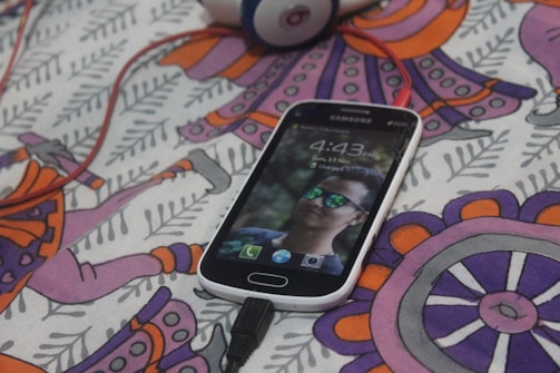 A Samsung smartphone displaying a lock screen with a person's face wearing reflective sunglasses. The phone is placed on a colorful patterned fabric with abstract designs in purple, orange, and pink shades. A pair of red headphones is partially visible in the background, connected to the phone.