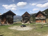 Three rustic wooden cabins are set against a picturesque mountain backdrop, surrounded by green grass and a few trees. The cabins have simple architecture with dark wooden exteriors, each with a distinct design. A fence made of logs encircles the area, adding to the rural charm of the scene. In the distance, snow-capped mountains and a clear blue sky with scattered clouds provide a tranquil atmosphere.