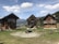 Three rustic wooden cabins are set against a picturesque mountain backdrop, surrounded by green grass and a few trees. The cabins have simple architecture with dark wooden exteriors, each with a distinct design. A fence made of logs encircles the area, adding to the rural charm of the scene. In the distance, snow-capped mountains and a clear blue sky with scattered clouds provide a tranquil atmosphere.