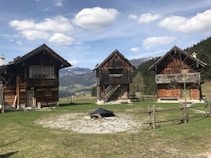 Three rustic wooden cabins are set against a picturesque mountain backdrop, surrounded by green grass and a few trees. The cabins have simple architecture with dark wooden exteriors, each with a distinct design. A fence made of logs encircles the area, adding to the rural charm of the scene. In the distance, snow-capped mountains and a clear blue sky with scattered clouds provide a tranquil atmosphere.