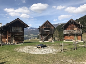 Three rustic wooden cabins are set against a picturesque mountain backdrop, surrounded by green grass and a few trees. The cabins have simple architecture with dark wooden exteriors, each with a distinct design. A fence made of logs encircles the area, adding to the rural charm of the scene. In the distance, snow-capped mountains and a clear blue sky with scattered clouds provide a tranquil atmosphere.