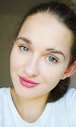 A close-up of a person with light skin, wearing a white top. The individual has green eyes and is smiling gently, offering a friendly, approachable appearance. Their hair is pulled back, and the lighting is bright and even.