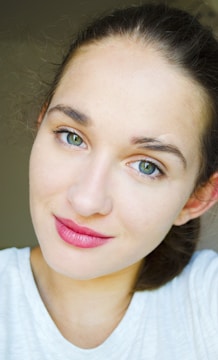 A close-up of a person with light skin, wearing a white top. The individual has green eyes and is smiling gently, offering a friendly, approachable appearance. Their hair is pulled back, and the lighting is bright and even.