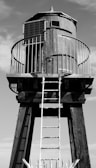 A tall wooden water tower structure with a cylindrical tank elevated on wooden stilts. A metal railing surrounds the tank, and a metal ladder leads up to it. There is a solar panel attached on one side. The image is in black and white with clear sky in the background.
