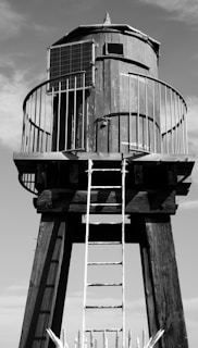 A tall wooden water tower structure with a cylindrical tank elevated on wooden stilts. A metal railing surrounds the tank, and a metal ladder leads up to it. There is a solar panel attached on one side. The image is in black and white with clear sky in the background.