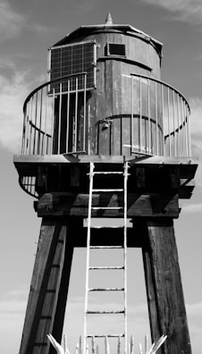A tall wooden water tower structure with a cylindrical tank elevated on wooden stilts. A metal railing surrounds the tank, and a metal ladder leads up to it. There is a solar panel attached on one side. The image is in black and white with clear sky in the background.