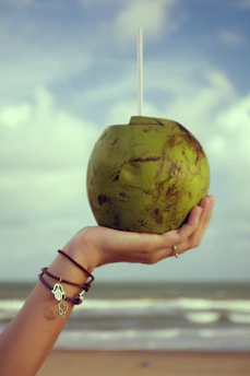 A hand adorned with bracelets holds a fresh green coconut with a straw against a backdrop of an overcast sky and a beach. The beach and ocean waves are slightly blurred, adding to the laid-back ambiance.