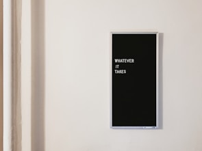 A minimalistic design featuring a framed black sign with the text 'WHATEVER IT TAKES' in white letters, mounted on a plain off-white wall. The composition suggests a motivational theme.