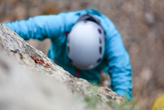 A person wearing a light blue jacket and a white helmet is climbing a rock wall. The focus is on the climbing gear, including a carabiner attached to the rock, suggesting an outdoor climbing activity.