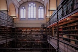 A multi-level library filled with rows of bookshelves, featuring tall arched windows that let in natural light. A central spiral staircase connects the different levels, and the architecture combines both classic and intricate details.