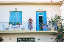An elderly woman is standing on an upper balcony, which features vibrant blue shutters and railings. The balcony is decorated with various plants and flowers in pots. The wall below the balcony displays a 'Primavera' sign for a restaurant that serves pizza and pasta.