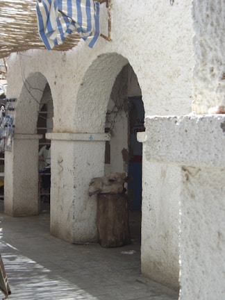 An exterior scene with arched stone structures and a woven roof, beneath which hangs a blue and white striped fabric. A severed animal head, possibly a cow or camel, is placed on a wooden post.