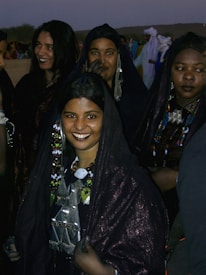 A group of women dressed in traditional dark attire, adorned with colorful bead necklaces and intricate jewelry. They are gathered closely together, with some sharing smiles. The background includes other people and desert scenery, suggesting a cultural or festive setting.