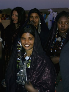 A group of women dressed in traditional dark attire, adorned with colorful bead necklaces and intricate jewelry. They are gathered closely together, with some sharing smiles. The background includes other people and desert scenery, suggesting a cultural or festive setting.