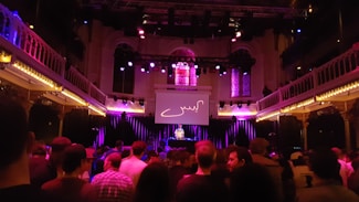 A concert venue filled with a crowd facing a stage with a DJ. The space features ornate architecture with balconies on both sides illuminated by purple and yellow lights. Stained glass windows are visible above the stage, and a large screen displaying an abstract design is positioned in the center.