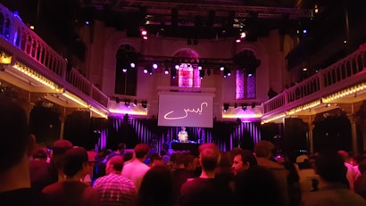 A concert venue filled with a crowd facing a stage with a DJ. The space features ornate architecture with balconies on both sides illuminated by purple and yellow lights. Stained glass windows are visible above the stage, and a large screen displaying an abstract design is positioned in the center.