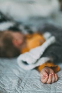 A child is sleeping or resting on a bed, partially covered with a gray and white blanket. The hand is slightly curled on the bed's surface, and the child's clothes are orange.