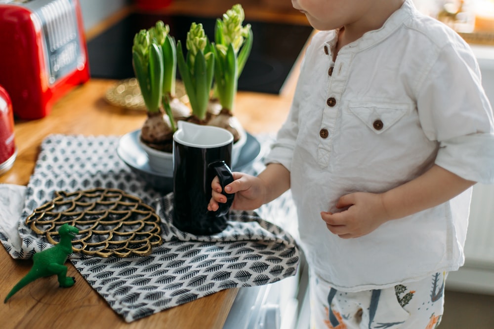 child holding black and white ceramic mug while standing near desk