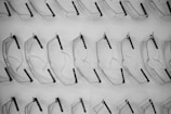 A pattern of clear safety glasses or goggles is arranged methodically on a white display surface. The lenses and frames are translucent, with black temple arms visible. The repetition and uniformity of the arrangement create a visually pleasing grid-like pattern.