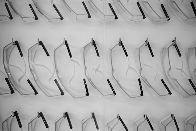 A pattern of clear safety glasses or goggles is arranged methodically on a white display surface. The lenses and frames are translucent, with black temple arms visible. The repetition and uniformity of the arrangement create a visually pleasing grid-like pattern.