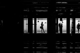 Silhouettes of people standing on a subway platform, with a bright backlight creating strong contrasts and casting shadows. The scene is framed by the dark structure of the subway station, highlighting the architectural layout and evoking a sense of motion or waiting.