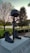 A military memorial featuring a helmet resting on a rifle that is pointed down, which is placed between a pair of combat boots. This arrangement sits upon a stone pedestal with text partially visible. The memorial is surrounded by a well-kept garden and small American flags. Trees and a body of water are visible in the background under a clear sky, suggesting a peaceful park-like setting.