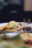 A close-up of a gourmet dish featuring a stuffed pastry topped with melted cheese, alongside a small portion of what appears to be a vegetable or grain salad. The background is softly blurred, emphasizing the dish.