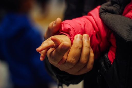 A close-up shot of a person's hand gently holding a child's hand. The child is wearing a bright pink jacket, and the adult's hand appears protective and caring.