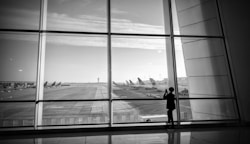 A black and white photograph of an airport runway viewed through large glass panels. Several airplanes are parked in the distance, and a solitary figure stands by the window, possibly taking a photograph.